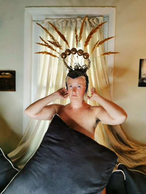 A white artist with blond hair wears a headpiece decorated with large golden wheat sheaves. A pillow covers her naked chest.