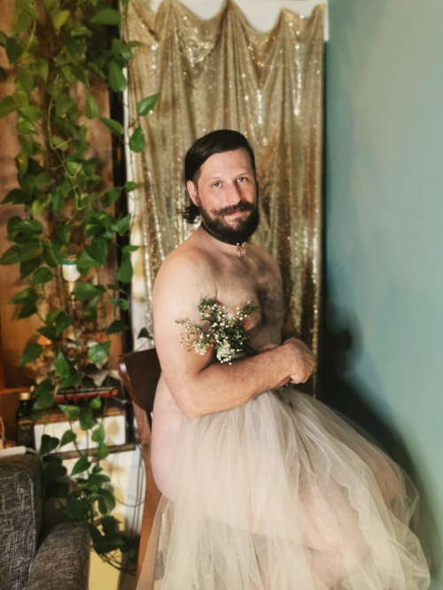 A white artist with long brown hair, mustache and beard sits in front of a gold background with a small bouquet of flowers and white tulle covering their chest and legs
