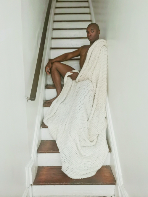 A dark-skinned artist with a shaved head sits on white and brown stairs, draped in a white blanket.