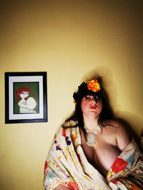 A light-skinned artist wearing a flowered headpiece, delicate necklace, and flowery fabric sits in front of a small painted portrait