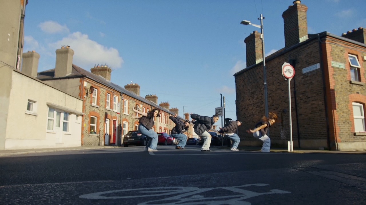 On an Irish street, five dancers wearing jeans and puffy jackets crouch low with their arms thrown back. 