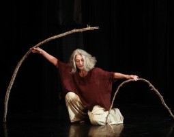 Dancer Merián Soto kneels with one knee slightly higher than the other. Her body faces the camera but her eyes are downcast. Her medium length gray hair falls towards her face. She is wearing a flowy rust-colored top and loose khaki-colored pants. In her outstretched hands she holds two curved branches. The branch in her left hand is nearly bent in half and almost touches the floor. The branch in her right hand extends in a graceful arc above her head, mirroring the casual grace in Soto’s pose.