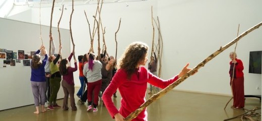 Dancers rehearse for the closing performance of Merián Soto’s exhibition last January celebrating her 25 years at Temple University. Marion Ramírez is center. Soto is to the right, overseeing the rehearsal and also participating. Each dancer is dressed in colorful, casual exercise clothing and holds a long branch vertically along their body, arms raised above their head and branch extended into the sky. From her vantage point, Soto is slightly removed; she is smiling.
