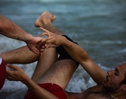"entangled" - With the ocean in the background, we only see the back of a man wearing a dirty blue shirt, James Barrett. He is reaching his hands behind him to touch another man, Miguel Alejandro Castillo, who wears red shorts and his lying on his back.
