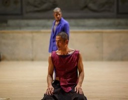 Movement Research at the Judson Church. In the foreground, Jungwoong Kim kneels. They are sitting back on their heels with their palms resting on their thighs. They wear black pants and a red sleeveless top. In the background of the church, another dancer, out of focus, walks behind the kneeling dancer. They wear a purple jacket. Neither dancer’s face is turned towards the camera; they both look away in opposite directions.