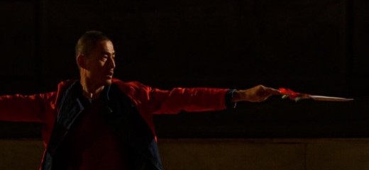 An artist stretches their arms side to side with their body positioned towards the camera. They are looking in the direction of one extended arm and appear to be holding a wand or blade in their hand.