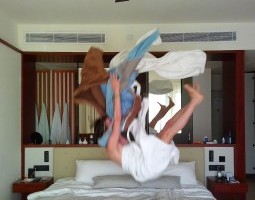 A blurred image of a body in motion and in the air. The body is wrapped in a white fabric (potentially a bathrobe) and bent in a V-shape as if falling onto the bed beneath. Tossed fabrics above them are brown, baby blue, grey, and white - possibly other clothes. The bed is made but rumpled and evokes a hotel room.