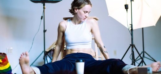 The performer, marisa illingworth, sits on the floor in a straddle with their body facing the camera. They are wearing a white crop top and navy sweat pants and have bare feet. A coffee cup sits in between their legs, white with a black lid, anonymous.Tattoos are visible on their arms. They look down and away from the camera. Behind them is photography lighting equipment; one of the lights is lit, putting the performer in the spotlight.