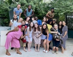 On a sunny day, a group of graduate students, and faculty huddled close together, smile at the camera. Their body language is loose and relaxed, and they look comfortable with one another.