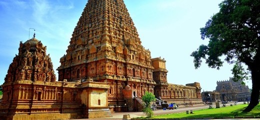 A view of the outside of the Brihadeeswara temple shows two large golden turrets with many intricate carvings, surrounded by a similarly carved wall. There is a bright blue sky behind the temple and a large tree on the other side of the image. 