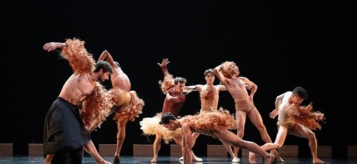 A group of performers on stage strike dynamic, expressive poses, adorned with elaborate red wigs and minimal costumes. Their movements and intricate formations evoke a sense of theatricality and playfulness, blending bold individuality with collective artistry. The dramatic lighting emphasizes the contrast between the performers and the dark background, heightening the visual impact.