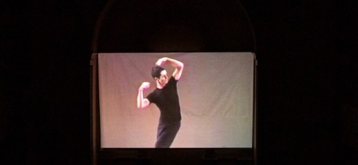 Against a white background Steve Paxton dances in black pants and a black t-shirt. He is leaning into his left hip, arching the left side of his body away from the hip, arms and hands loosely in the air around his head.