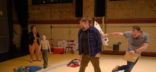 Alex Torra winces as Benjamin Camp hits him on the back with a patterned pillow. Benjamin is jumping with the force of his throw, clutching a clipboard and wearing a stopwatch around his neck. Sydney Camp, Benjamin’s daughter, calmly walks between a basket of toys and a pile of clothes behind them. Rachel Camp, Benjamin’s sister, watches behind Sydney, a hand in front of her mouth hiding an amused smile.