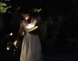 A young Black woman (Arianna Polite) holds a wooden bowl glowing with warm white light. She looks downward, sprinkling marbles of light onto the ground as darkness falls after sunset.