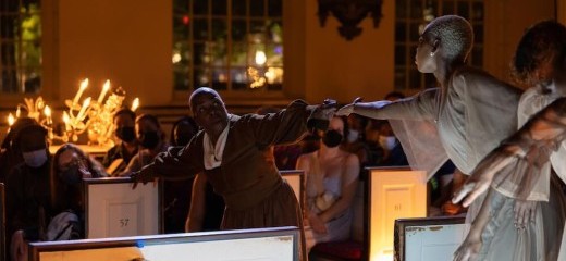 A mature Black woman (Rhonda Moore) stands in the center of two rows of pews with her arms outstretched. She grips the side of one pew reaching towards the open palm of another Black woman (Song Aziza Tucker) who stands in George Washington’s pew at Christ Church.