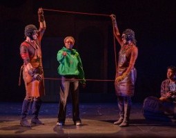 In a vast black expanse with a dark brown floor, two Gogglers in warm colored shirts, leather aprons, and steampunk goggles hold up a square of red thread around Temple, a Black girl in a bright green sweatshirt. She's pointing at the viewer as her brother, Arturo, looks on in interest while sitting on the ground.