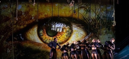 he image features glowing amber eyes projected onto the backdrop of the stage. On the sun-dappled-like stage, a group of BalletX dancers stands tight together, mirroring the character Jack’s command to lift their strong and angular arms to their hearts. Dressed in black, slim-cropped jackets and shorts, the dancers’ shrugged shoulders convey a sense of suspense or reverence. The atmospheric lighting, combined with the haunting gaze of the eyes, creates a mood of mystery and psychological intensity, reinforcing the ballet’s themes of surveillance, power, and primal instinct.