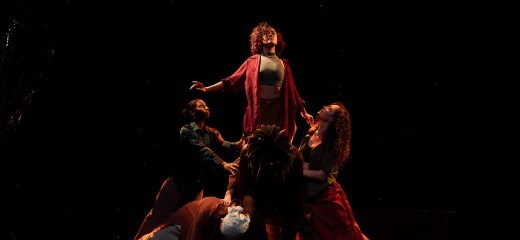 A dancer stands atop a group of other dancers, expanding their chest and focus upwards with an uplifted energy. They wear a maroon button-up shirt and don curly grown hair. The other dancers bend in towards this dancer, supporting and lifting them up. They wear multicolored pedestrian clothing and are lit by a warm, dim, light. They are soft with energy that extends upwards.