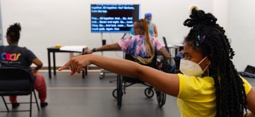 How We Move artists rehearsing majorette dance in the studio. Featured are Hector Machado, Jackie Robinson, and Kayla Hamilton. Everyone is masked with their left arm and index finger pointing outward to the side, and their right hand on their hip. The shot is taken from behind the dancers. In the front, captions are being shared on a large TV screen.