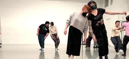 In a bright white room with light grey marley, dancers are clustered to the right side of the image in pairs, sharing weight. Two pairs of dancers are connected at the shoulders, one pair is connected by the hands, and we can see the legs of two hidden dancers standing back-to-back. There is an implication of motion, through shadows, blurred limbs, and raised feet.