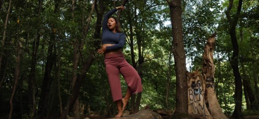 Balanced on a fallen tree in a wooded area a dancer poses wearing maroon pants and a blue long sleeved crop top. She balances on her right foot, touching her pointed left foot against the right ankle. Her left hand reaches across her midsection with the hand and fingers spread wide. Her right arm reaches up over her head, fingers spread wide, palm down. She softly gazes down, her mouth relaxed open.