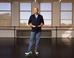 Christopher Kaui Morgan gazes off to the side, smiling serenely in a studio that radiates warmth. Outfitted in various shades of blue, his hands rest gently on his hips in the pockets of a navy cardigan, as his weight shifts over to one leg. He is an image of confident grace, wearing chunky high-heeled boots that add a splash of exuberance to his professional demeanor.