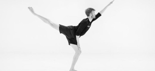 A young man facing the right, wearing black shorts and matching black t-shirt, beautifully extends his left arm forward and right leg back.