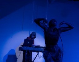 Vyette Tiya (she/her) smiles with her eyes closed and head tilted upwards, arms lofting upwards as she descends downwards. She wears baggy pants and a textured crop top, and is washed in a dark blue light. Two white chains hang in the background above her head. To her left is Daniel Sohn with a mixing board and laptop, focused on mixing for his music. He is wearing a baseball cap, t-shirt, and pants.