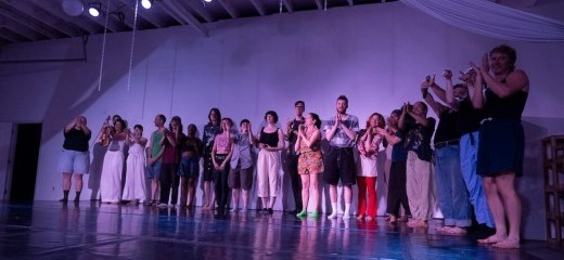 Full cast clapping onstage with the house lights up in a blue and purple light, taking from a low angle shot to their right.