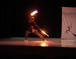 Yuki Ishiguro holds two glowing sticks in either hand. Wearing a loose-flowing silk top, he stands in power, pointing one stick to the ground, the other held above his head.