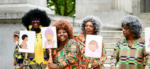 Standing in front of an old building with stone steps, four Black women of various ages stand wearing vivid shirts with complex patterns. All donning afros, three of the women hold up signs which show hand-drawn illustrations of infants or children, while the fourth has her hands resting on her walker. Two of the women smile radiantly.