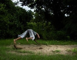 Marguerite Hemmings is surrounded by green trees and grass, pictured mid-air, as if descending to the ground head-first. One arm is outstretched to break the fall while the other arm is bent around their head, and their feet have kicked up the sand behind them.