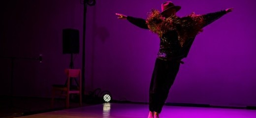Bathed in purple light, Adam Kerbel leans to one side with arms spread, a gold fringe flying from his shoulders as if he just landed a jump. His face is hidden by shadow and a large brown fedora.