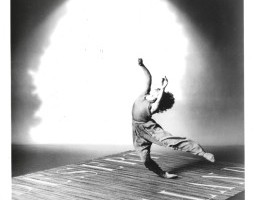 It’s 1986. Lisa Kraus, positioned slightly to the right of center of the image, falls to the side over one ankle, the other leg outstretched behind her. She raises her arms upward and lifts her head back, hair flying behind her. She dances in a spotlight on a mat on the floor that reads “DESERT ISLAND” in all capital letters. The image, captured by acclaimed dance photographer Lois Greenfield, is in black and white.