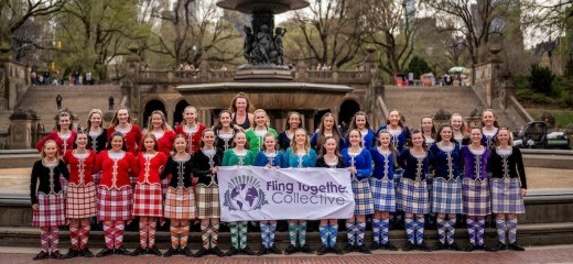 Two rows of Highland dancers stand in front of the Bethesda Fountain at one of the entrances to Central Park in NYC. There are trees in the background. The dancers are clad in bright plaids and knee socks in orange, red, green, blue, and purple. A few in the first row hold a sign that says Fling Together Collective