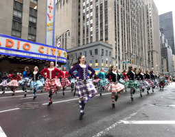 On the Avenue in front of the red sign above Radio City Music Hall, a group of Highland dancers are pictured landing out of a leap onto their right toes, the left leg bent at the knee out to the side, with the left foot touching the opposite leg just below the knee. They have their hands on their hips and broad, bright smiles. The brightly colored white and red, dark blue, light blue, purple, and green plaids of their Tartans and knee socks stand out on the cloudy day.