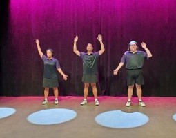 pink backlight, three dancers, left most has right arm up and left arm down, middle has both arms bent upward, right has bent left arm and straight right arm, all in purple polos, two white set pieces frame the image with black curved lines.