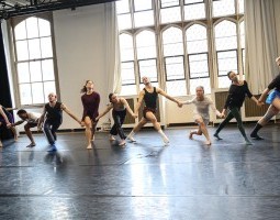 A line of eleven people are holding hands in front of bright large windows, their legs lunging and their heads turned in different directions. They were sweat pants and t-shirts, shorts or tank tops, all of various colors. Some of them are looking to their right and down, others looking up and left.