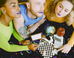 three people wearing brightly colored clothing and blue lipstick hover closely together, looking off into the distance. they are leaning over miniature figurines of a desktop computer, a black and white checkered floor, a globe and other tiny toy objects.