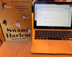 A copy of The Swans of Harlem stands next to an open laptop. The cover of the book is gold, with black and white photographs of five Black ballerinas in retiré, leaping, and posing in arabesque and with a pointed foot. The book’s subtitle is also visible on the cover: Five Black Ballerinas, Fifty Years of Sisterhood, and Their Reclamation of a Groundbreaking History.