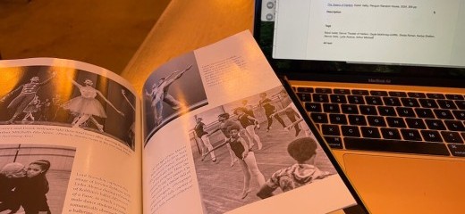 A copy of The Swans of Harlem is open on a table next to an open laptop. The book is open to a photo spread. The black-and-white images depict dancers from the early days of Dance Theatre of Harlem, including an image of Lydia Abarca being coached in Afternoon of a Faun, final bows, Gayle McKinney-Griffith being partnered in arabesque, and a photo of Marcia Sells in class at ten years old. Sells is jumping in second position with her feet pointed and arms outstretched, looking down towards her left hand.