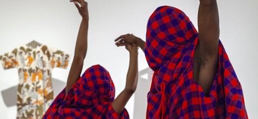 Two figures entwine in a tender embrace, their bodies wrapped in rich layers of Maasai Shuka, hands poised with balletic grace—an intimate tableau of support, connection, and quiet strength.