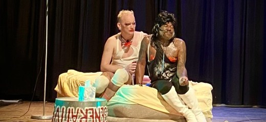 Two queer people sit on an inflatable bed. A metal basin flipped upside down is used as a table.