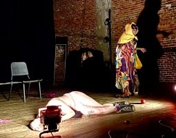 A white gay man lies on the floor, almost fully naked. A Black gay man dressed in a tropical dress with a hair scarf, sunglasses, and a floral bag stands next to them, extending their arms as if asking, “What’s the matter?”
