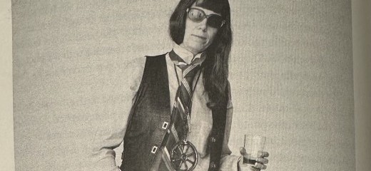 A photo of a page in the book. A black and white photo of Jill Johnston. She has long hair, aviator sunglasses, and is wearing a white long sleeve button up, a black vest, and a wide striped tie. The photo is cut off at her hips. She leans into her left hip and has her right hand in her pocket. A glass in her left hand.