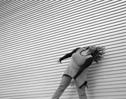 A black & white image of Kayla Hamilton, a dark-skinned Black woman. She is dancing in front of a textured wall that has horizontal layers. Her arms are energetically reaching down to the diagonal as her head is tilting to the diagonal with her dreads moving back in that same direction. She wears a diagonally striped long sleeve shirt with pants. Photo by Travis Magee.