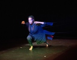Danielle Currica, one of the four soapbox dancers, lunges low and extended on a diagonal to the left front of the stage. She looks intently, perhaps searching, with wide eyes, wearing a long blue garment with fabric extensions behind her attached to four ropes, faintly seen extending into darkness beyond.