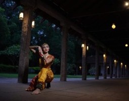 Lakshmi Thiagarajan, author of the article, kneels gracefully on the ground in a covered walkway outdoors, lit by lanterns. One leg is forward, clad in yellow and red silk pants with a percussive bracelet on her ankle. Her clasped hands frame her face, adorned with a bindi and golden headband. She looks off to the side with a small smile.