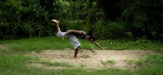 Marguerite Hemmings stands on one leg in active posture with one leg stretched behind them, arms outstretched in opposite directions, and torso parallel to the ground. They are surrounded by green trees and grass, standing on a patch of sand.