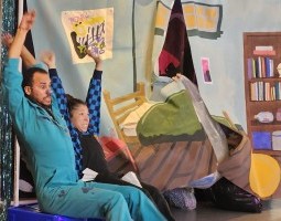 Two people dressed in blue sit on top of an overturned recycling bin, with their arms extended overhead as if riding a rollercoaster. They have nervous looking facial expressions, and are in front of a curtain backdrop which is painted like a bedroom. There is a hand coming from behind the backdrop, grasping it, as if preparing to enter the stage.